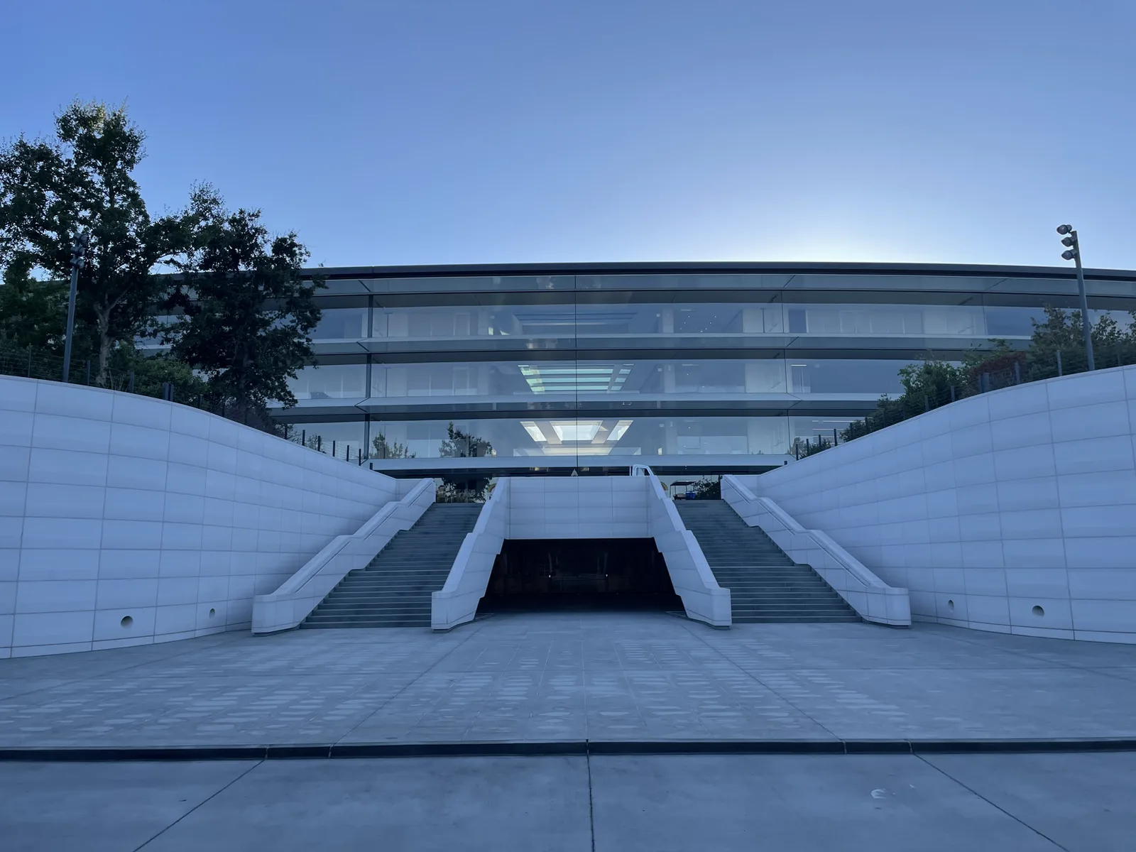 Twin concrete stairs rising either side of a sunken entry, flanked by long sweeping pale walls beneath a glazed facade