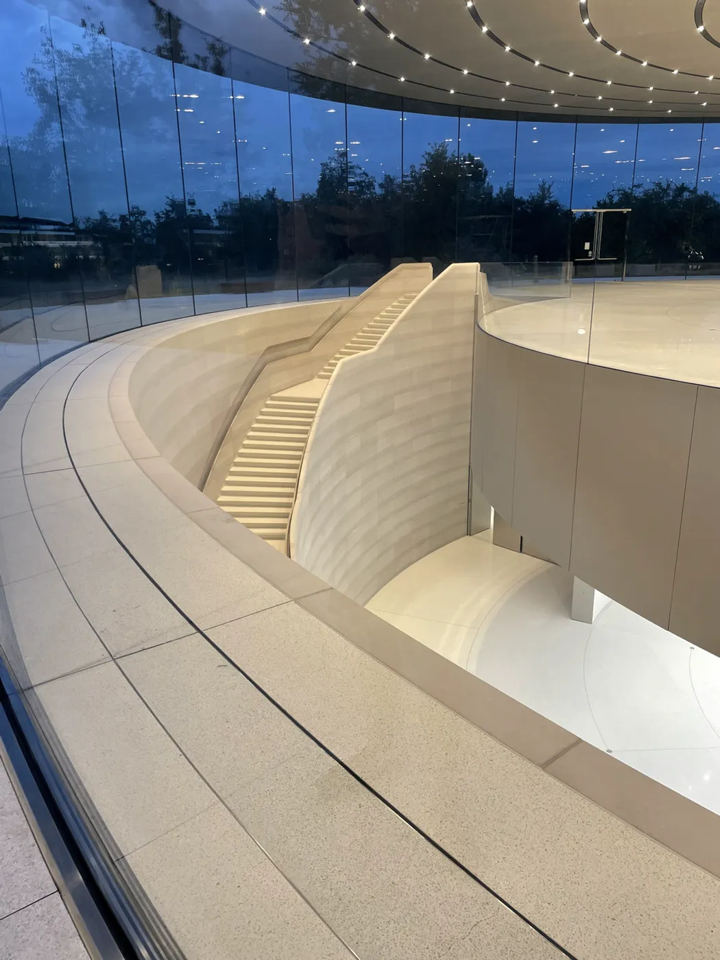 A curved stair descending beside the theatre&rsquo;s glass drum, ringed terrace walls stepping around it