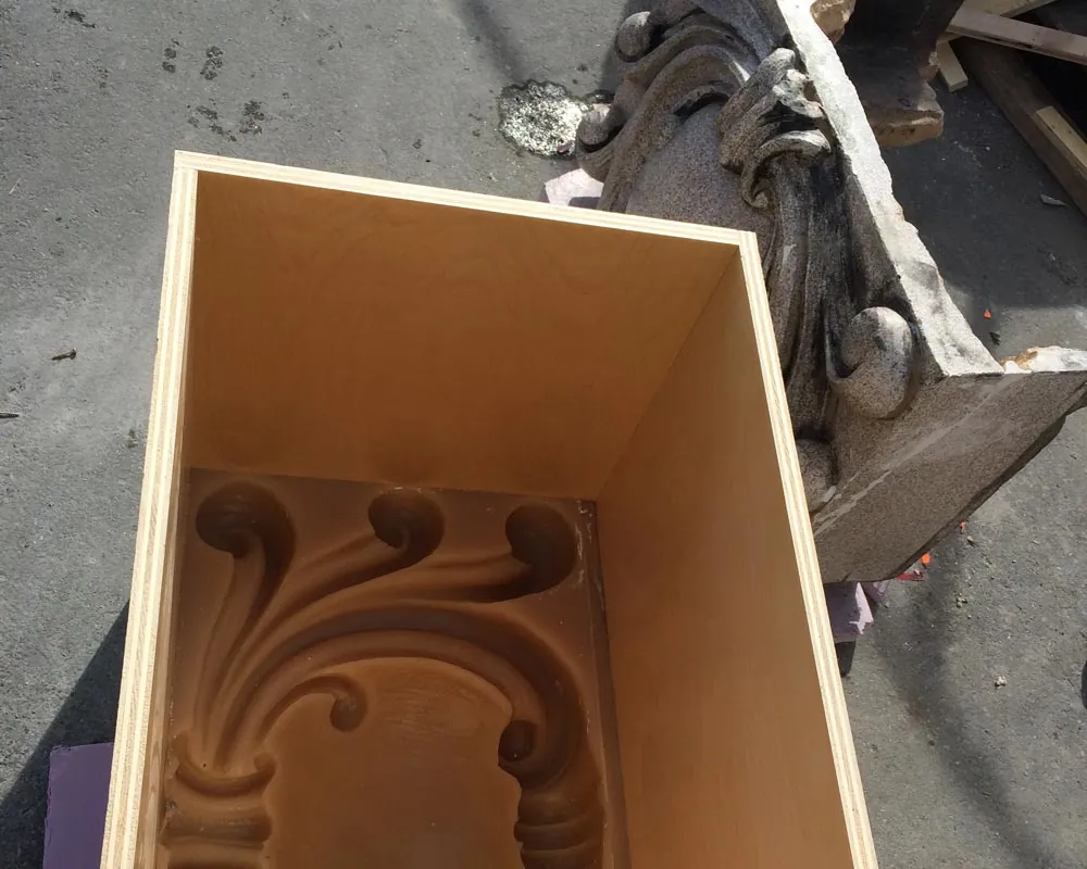 A rubber mold of an ornate scroll profile in its wood crate, beside the weathered original terracotta block