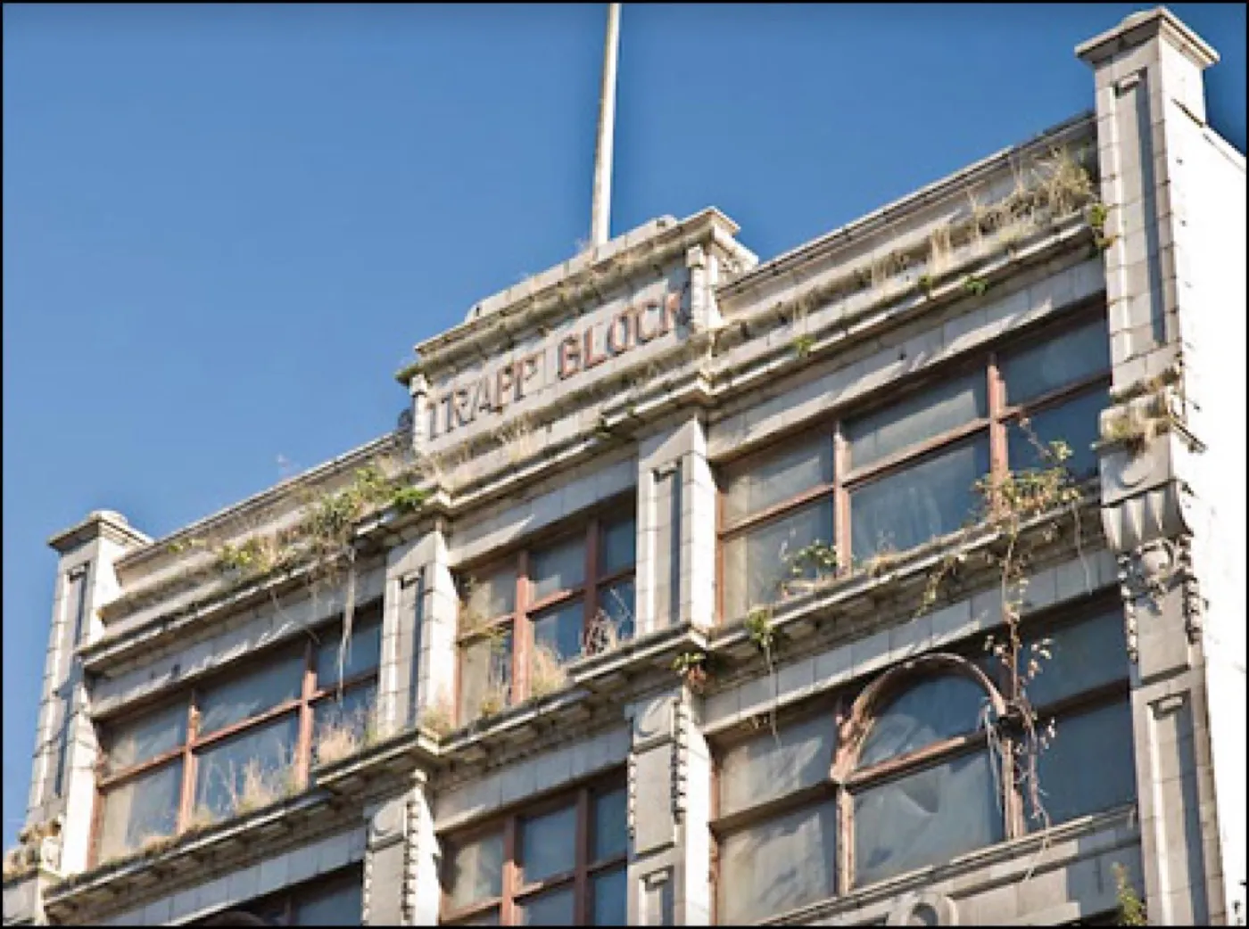 The derelict Trapp Block facade against blue sky, its carved signage weathered and plants rooted in the cornices