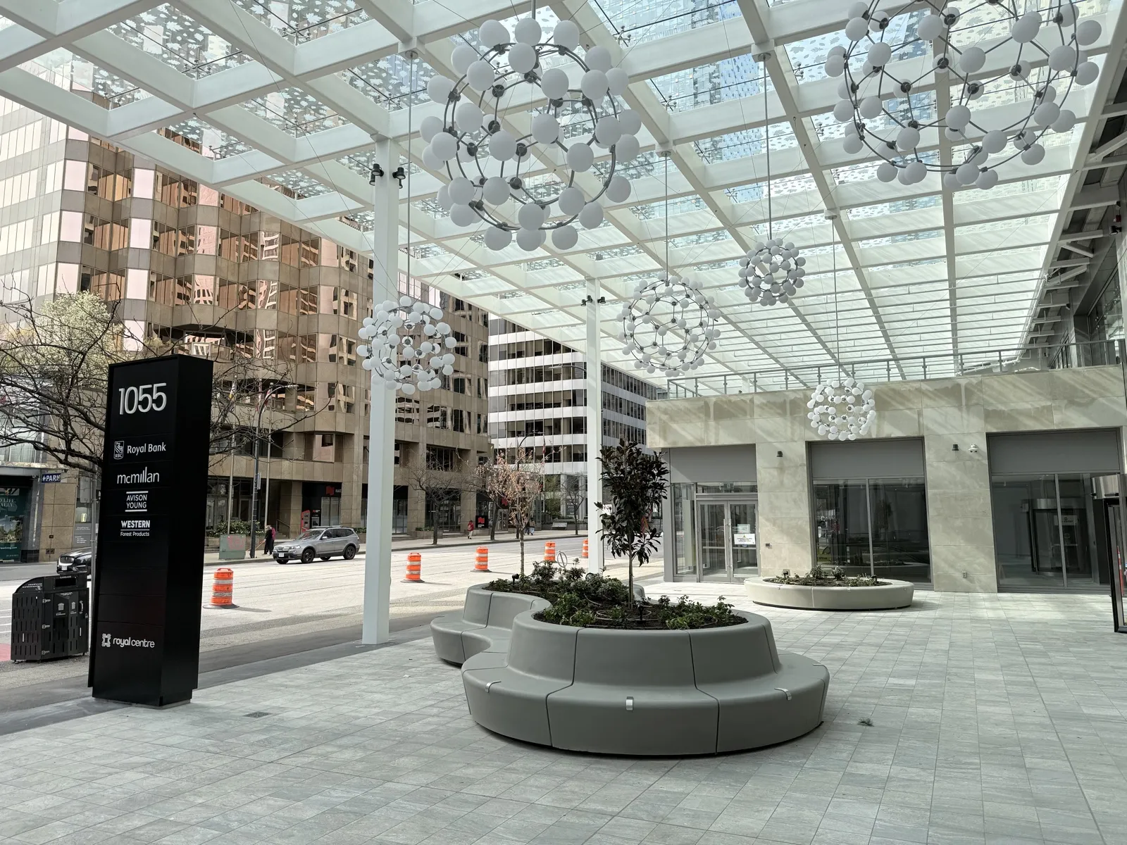 The Royal Centre plaza under its new glass canopy — a precast seating ring wrapping a planted island beneath orb chandeliers