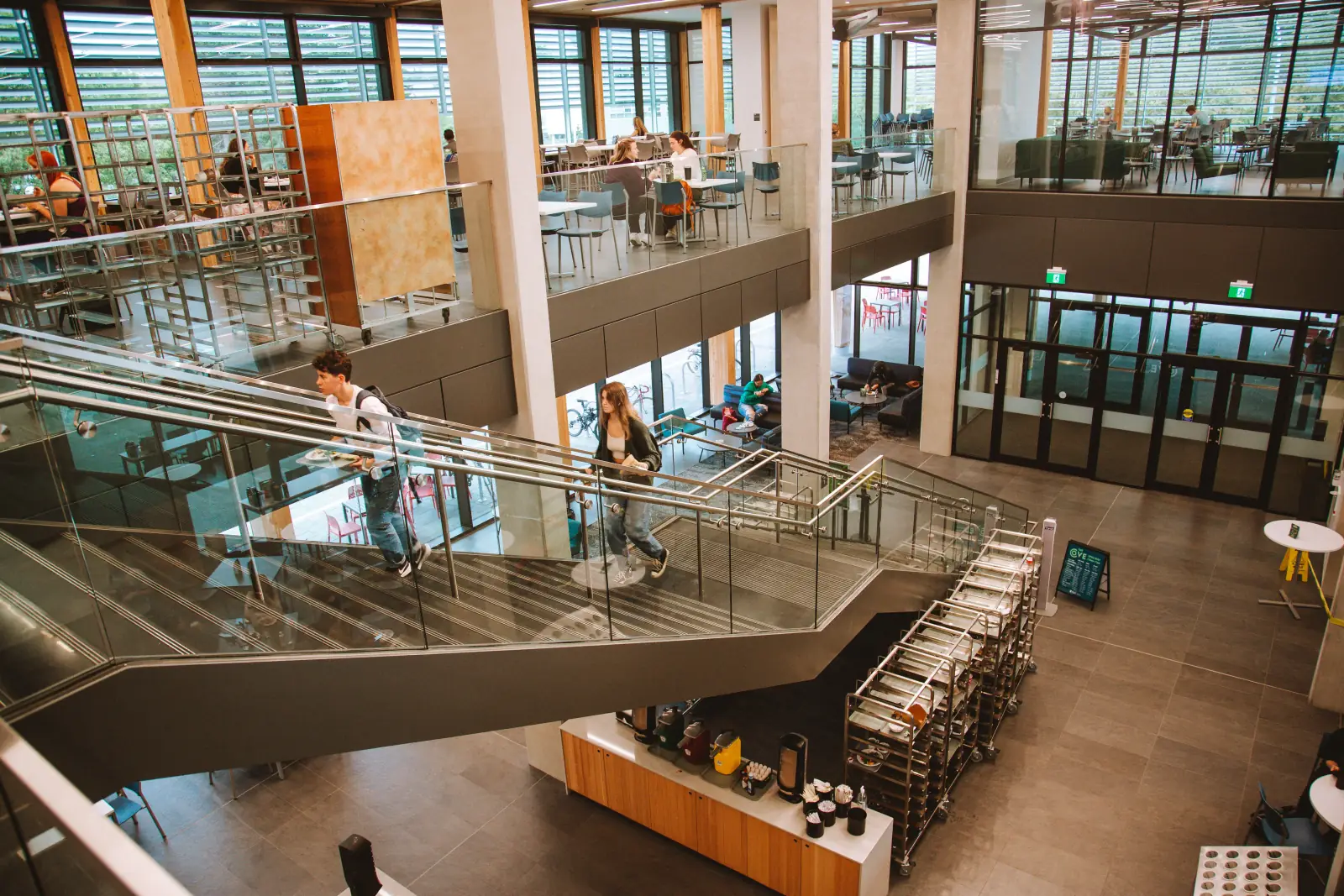 The UVic dining hall with a precast UHPC feature stair crossing the room, students moving through beneath a mass-timber ceiling